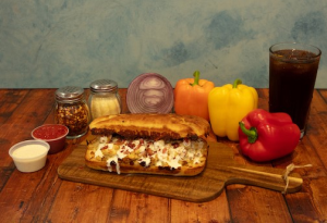 Genos Giant Slice sandwich on wooden board with peppers, condiments, and drink on rustic table