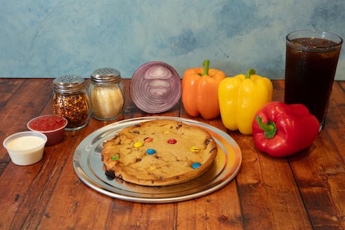 Genos Giant Slice pizza with M&M toppings on metal plate, surrounded by peppers, condiments, and soda on wooden table