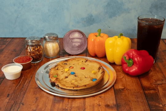 Genos Giant Slice pizza on metal plate with toppings, surrounded by peppers, condiments, and drink on wooden table