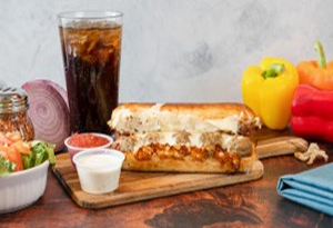 Genos Giant Slice sandwich on wooden board with side dishes and soda drink