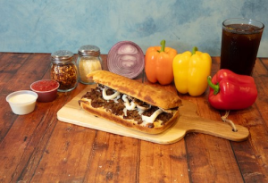 Genos Giant Slice sandwich on wooden board with peppers, condiments, and drink on rustic table