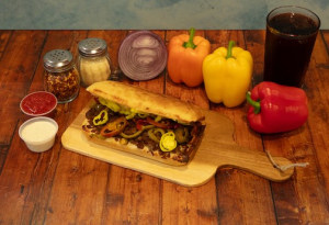 Genos Giant Slice sandwich on wooden board with peppers, condiments, and drink beside it