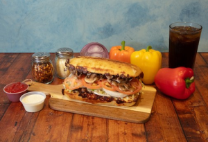 Genos Giant Slice sandwich on wooden board with peppers, condiments, and soda on rustic table