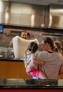 Genos Giant Slice pizzeria: family with child holding pizza dough at counter, chef smiling behind stainless steel kitchen equipment