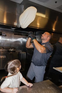 Pizza chef tossing dough in kitchen, child watching nearby, stainless steel appliances visible