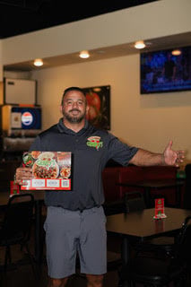 Man in gray shirt holding Genos Giant Slice pizza box, gesturing in pizzeria with tables and TV in background